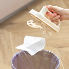 Hand using a white plastic bag holder to extract a white plastic bag from a clear trash bin on a wooden floor.