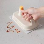 Hand using a white cleaning tool with a yellow handle to wipe up a brown stain on a light surface.